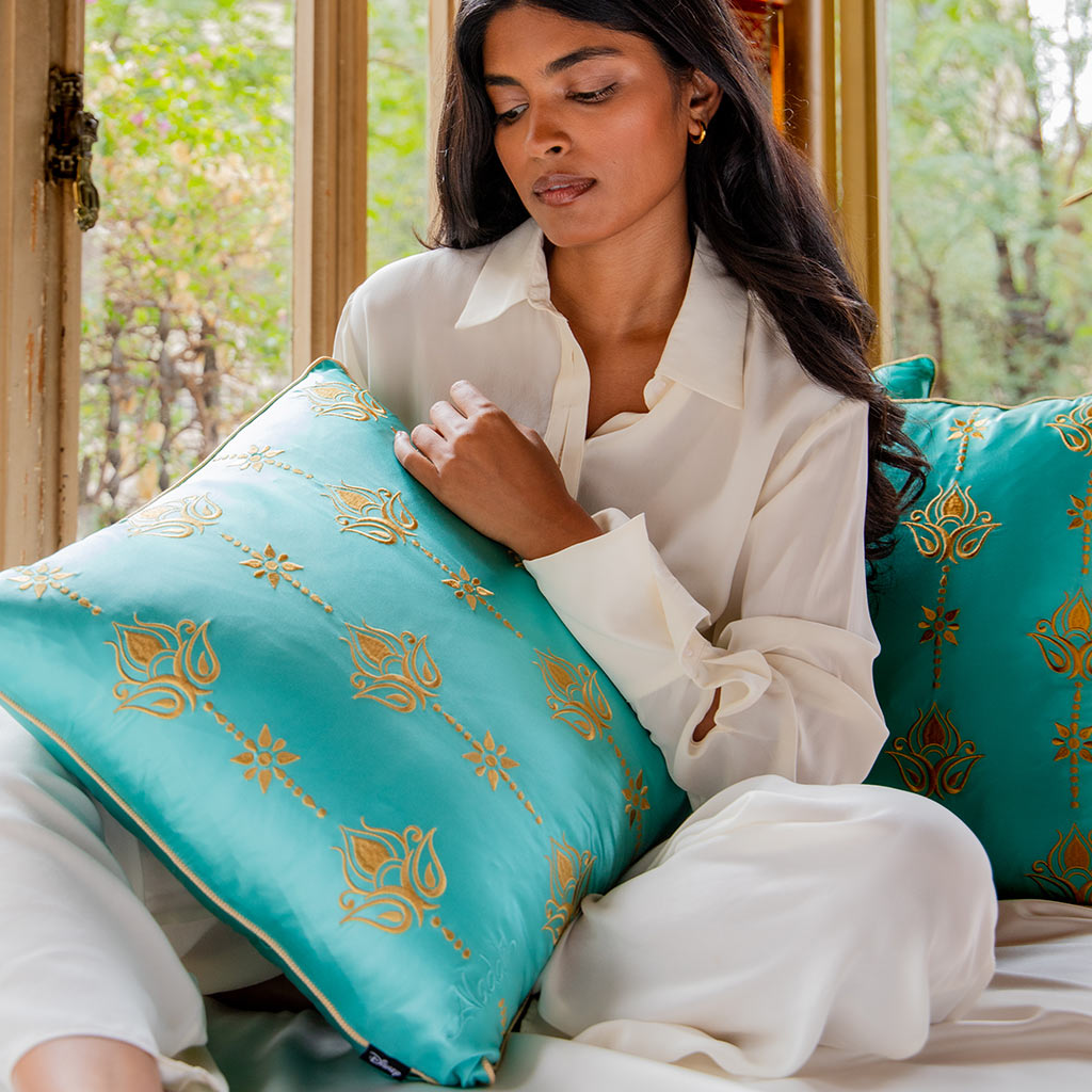 A woman in white sits by a window holding drowsysleepco's Jasmine Cushion Cover—turquoise silk with gold patterns. A matching pillow rests beside her, with green trees visible outside.