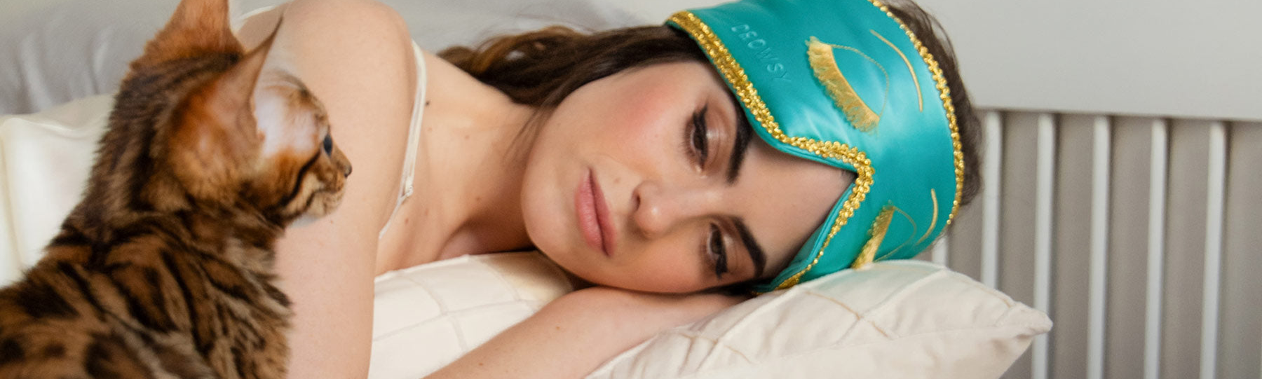 A woman wearing a turquoise sleep mask with gold trim rests on a pillow, eyes closed, while a brown-striped cat sits nearby, looking at her.