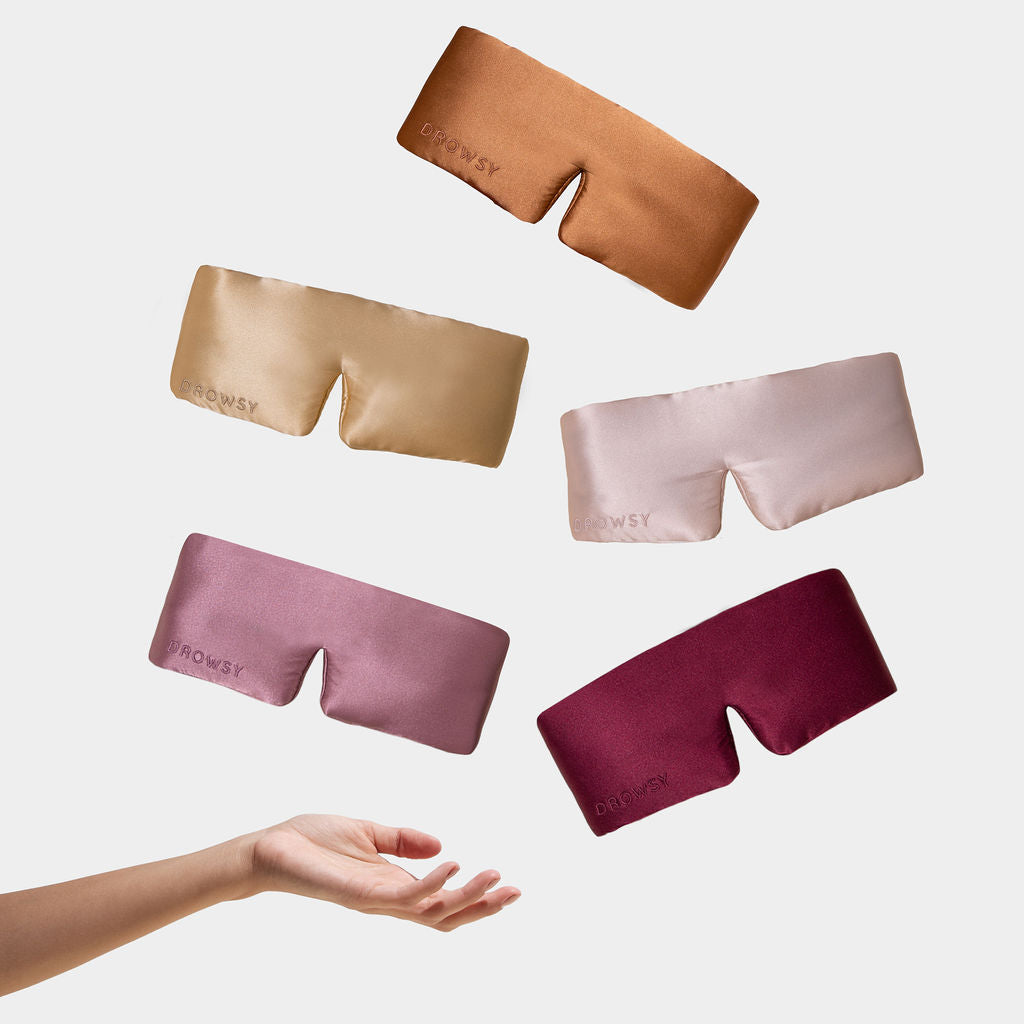 A hand reaches up toward five floating drowsysleepco Family Pack silk masks in tan, gold, light pink, mauve, and burgundy—each labeled “DROWSY”—against a white background, offering the ultimate blackout eye mask for beauty sleep.