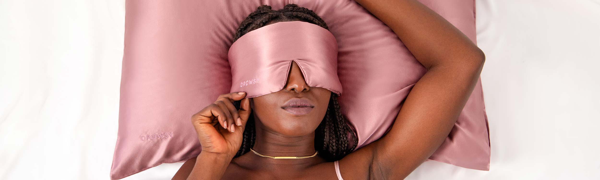 A woman lying on a pink satin pillow wears a matching pink satin sleep mask, with one hand gently touching the mask. She has braided hair and wears a delicate gold necklace.