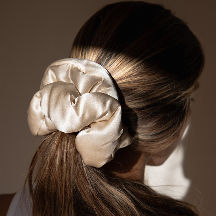 A woman with brown hair wears the Rajah Padded Scrunchie by drowsysleepco in shiny beige satin, securing a low ponytail. Soft light highlights the scrunchie and her hair, with a softly blurred background.