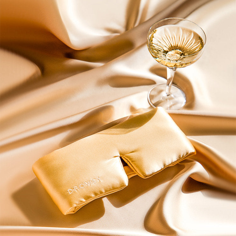 A satin eye mask with DR. OYSHI embroidered on it lies on silky gold fabric beside a glass of white wine, creating a luxurious and relaxing scene.