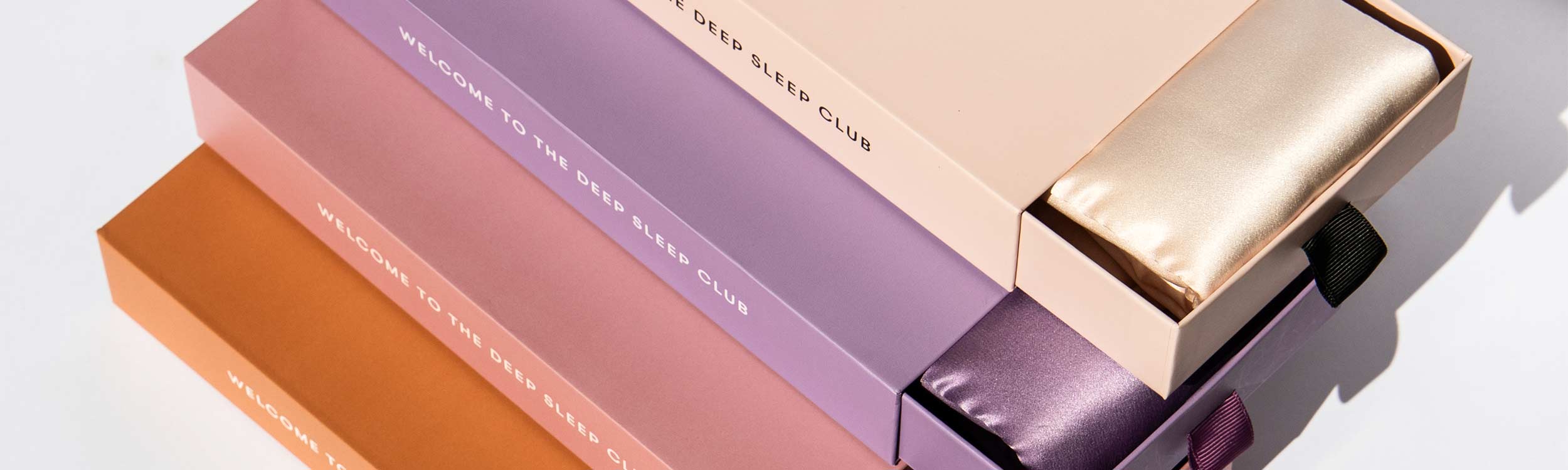 Four rectangular boxes in pastel shades—peach, lavender, gold, and orange—stacked diagonally. Two boxes are partially open, revealing shiny satin sleep masks inside. Text on boxes reads “WELCOME TO THE DEEP SLEEP CLUB.”.