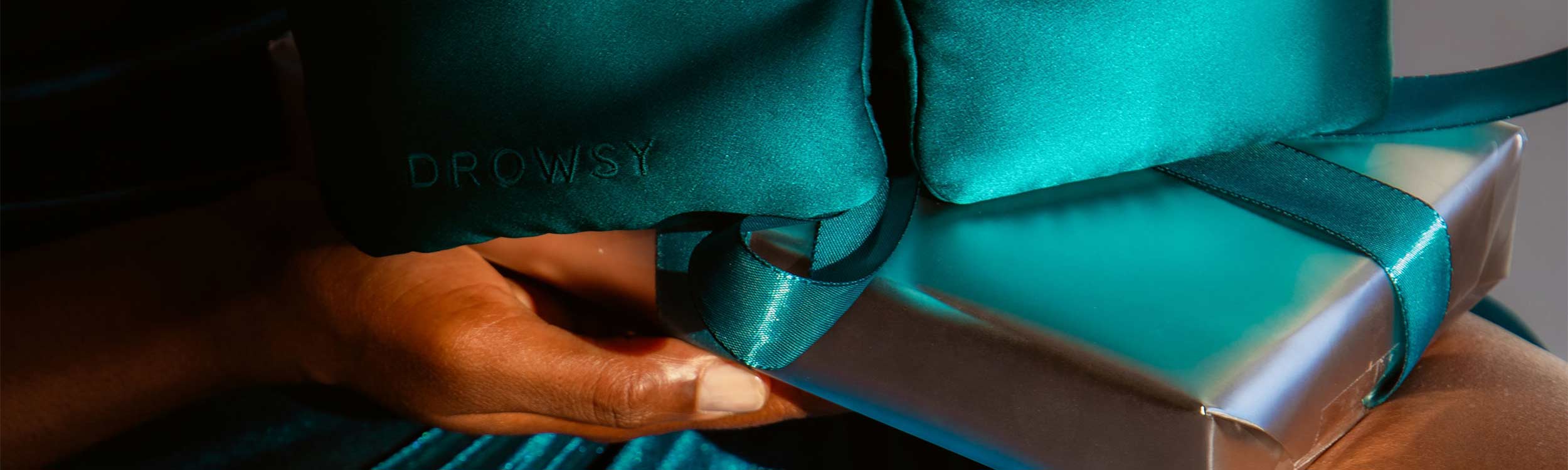 A hand holds a shiny, silver gift box wrapped with a teal ribbon. On top of the box is a teal satin eye mask labeled DROWSY, with fabric matching the ribbon.