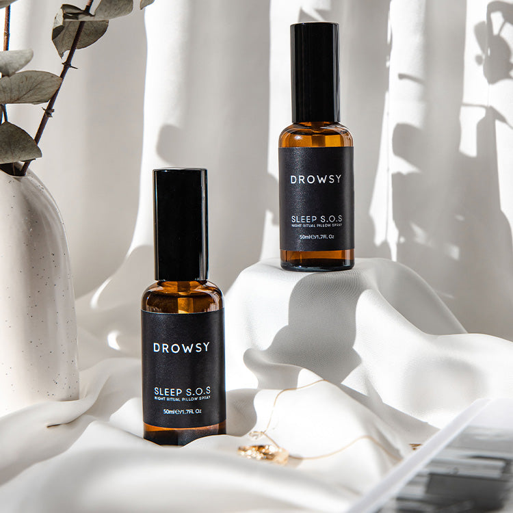 Two amber glass spray bottles labeled Drowsy Sleep S.O.S sit on a white, draped fabric surface with soft shadows. Eucalyptus leaves and a gold necklace are partially visible nearby.