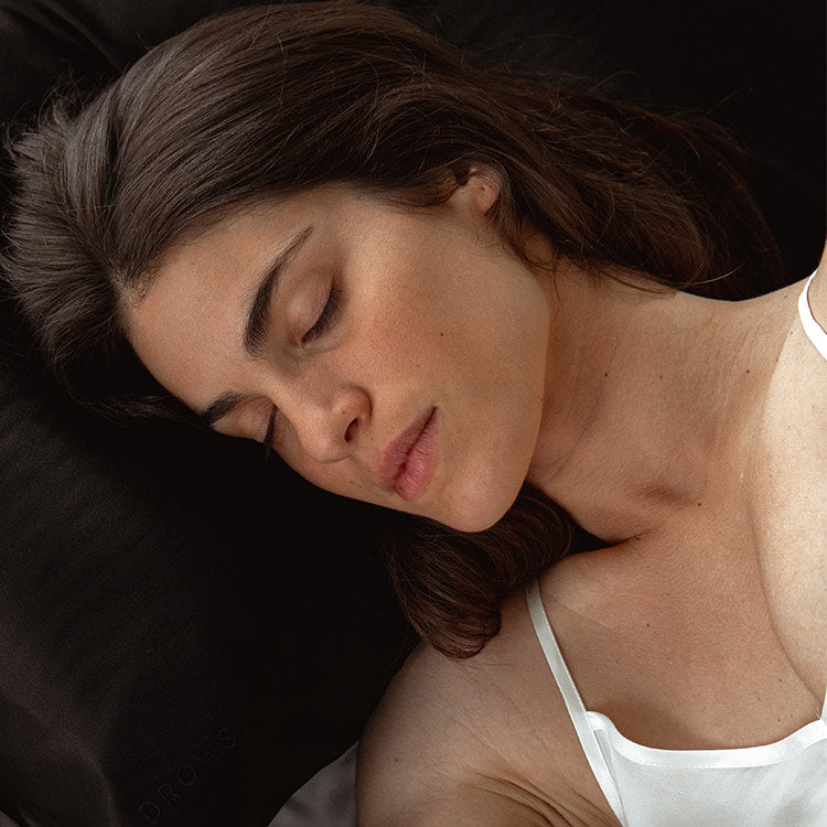 A woman with long brown hair is sleeping on her side, resting her head on a black pillow. She is wearing a white tank top and appears calm and relaxed.