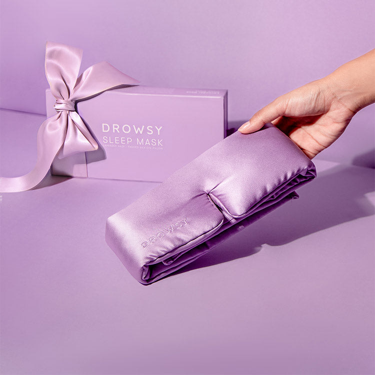 A hand holds a shiny lavender sleep mask in front of a matching purple Drowsy Sleep Mask box with a large satin bow, all set against a pastel purple background.