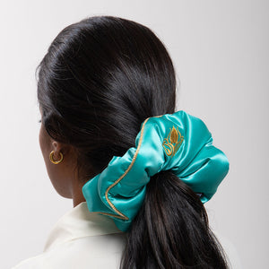 A woman with dark hair in a low ponytail, secured by the drowsysleepco Jasmine Padded Scrunchie in turquoise and gold, wears gold hoop earrings and a white top. She's photographed from behind against a light gray background.
