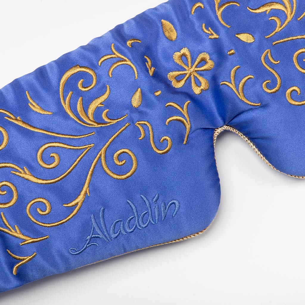 A close-up of drowsysleepco’s Magic Carpet Sleep Mask, a blue mulberry silk mask featuring gold embroidered swirls and “Aladdin” stitched in blue—designed as a luxurious blackout sleep accessory for restful nights.