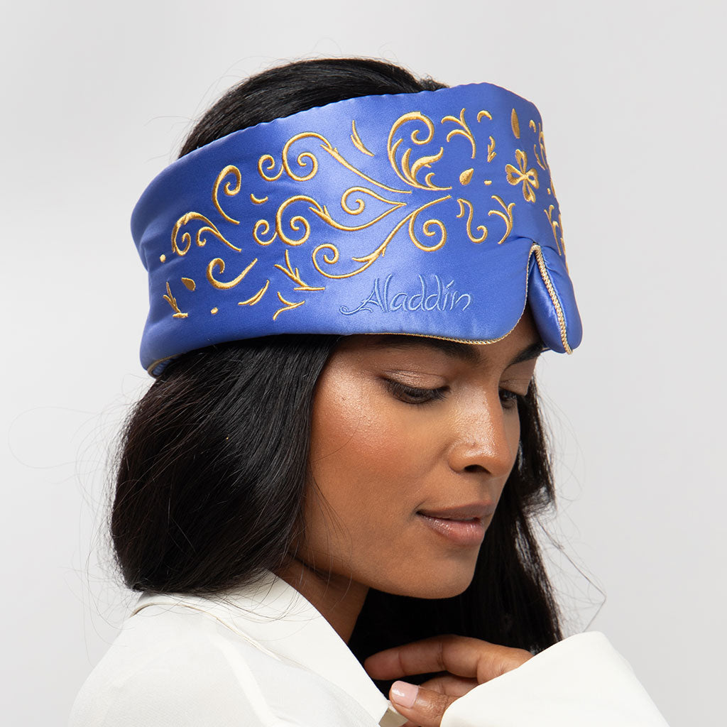 A woman with long dark hair wears a royal blue headband embroidered with gold swirls and "Aladdin," looking down in a white top while holding the drowsysleepco Magic Carpet Sleep Mask against a plain light background.