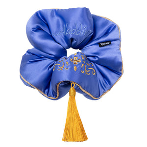The Magic Carpet Padded Scrunchie by drowsysleepco is a royal blue oversized silk scrunchie with gold embroidery, a gold tassel, "Aladdin" stitched on it, and a Disney tag—designed to boost hair thickness with a magical touch.