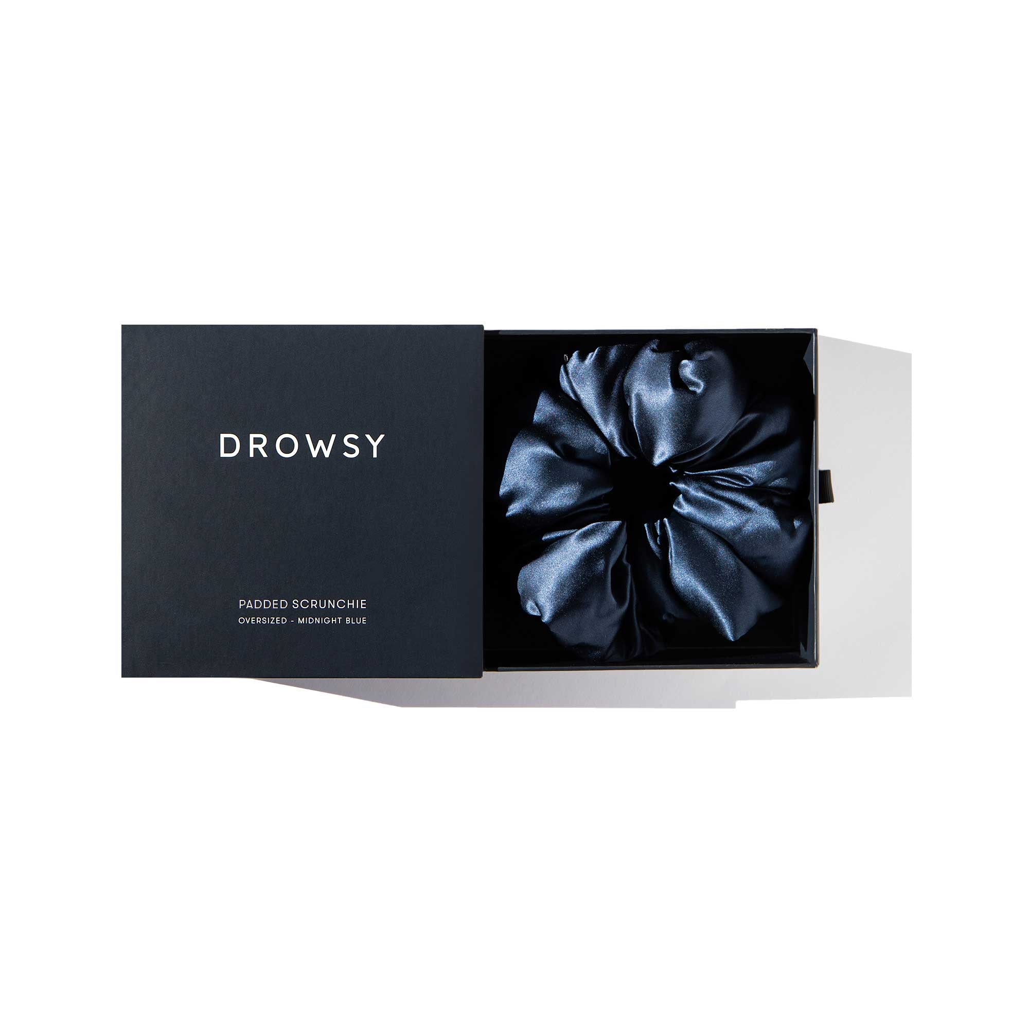 A Midnight Blue - Padded Scrunchie by drowsysleepco sits inside a sleek black branded box, its lid slightly open to reveal the oversized, padded silk hair accessory designed for added volume.
