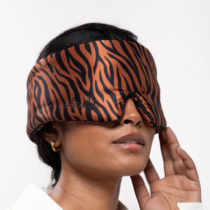 A woman wears the drowsysleepco Rajah Sleep Mask, featuring brown and black tiger stripes in mulberry silk, covering her eyes and part of her nose. She gently touches the mask, accessorized with gold hoop earrings and a white collared shirt.