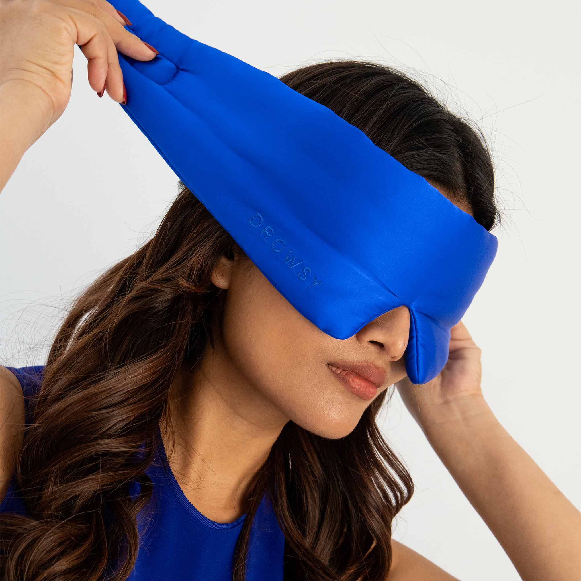 A woman with long brown hair wears a bright blue eye mask, adjusting it over her eyes with one hand. She is dressed in a matching blue top against a plain light background.