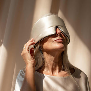 A woman with long gray hair, relaxed in soft natural light, wears drowsysleepcos Dusty Gold - Pipe Dreams Edition mulberry silk sleep mask and matching top, gently touching the luxurious mask.