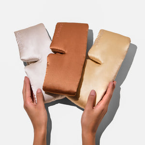 Two hands hold drowsysleepco’s Besties Pack: three luxury mulberry silk sleep masks in cream, bronze, and gold with “DROWSY” elegantly embroidered on the front, all arranged side by side on a white background.