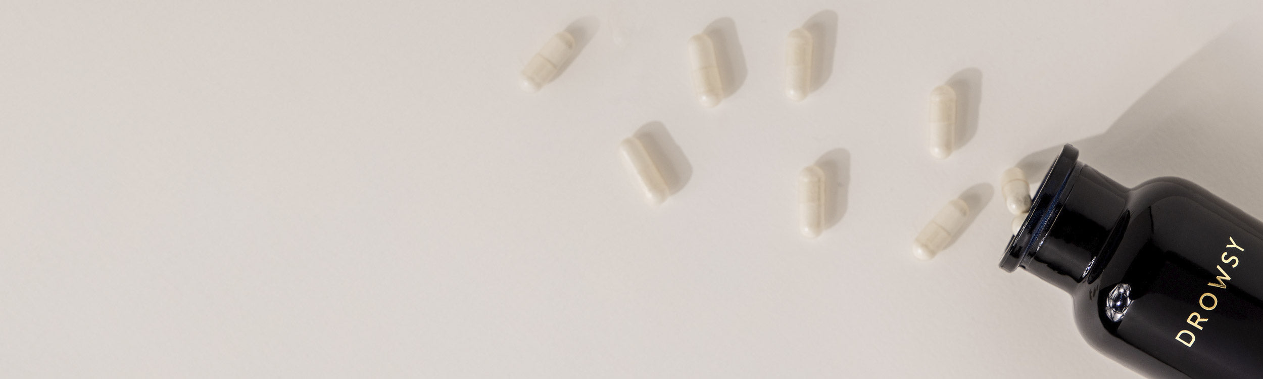 A black bottle labeled DROWSY lies on its side with white capsules spilling out onto a light beige surface.