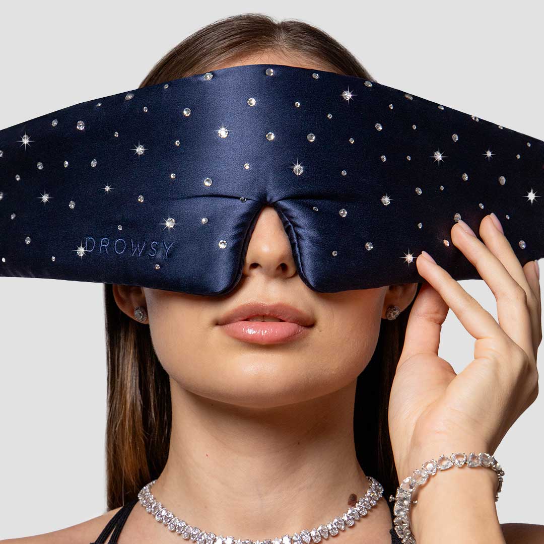 A woman wearing a navy blue, star-patterned sleep mask that covers her eyes and part of her nose. She holds the mask with one hand and wears sparkly jewelry, including a necklace and bracelet.