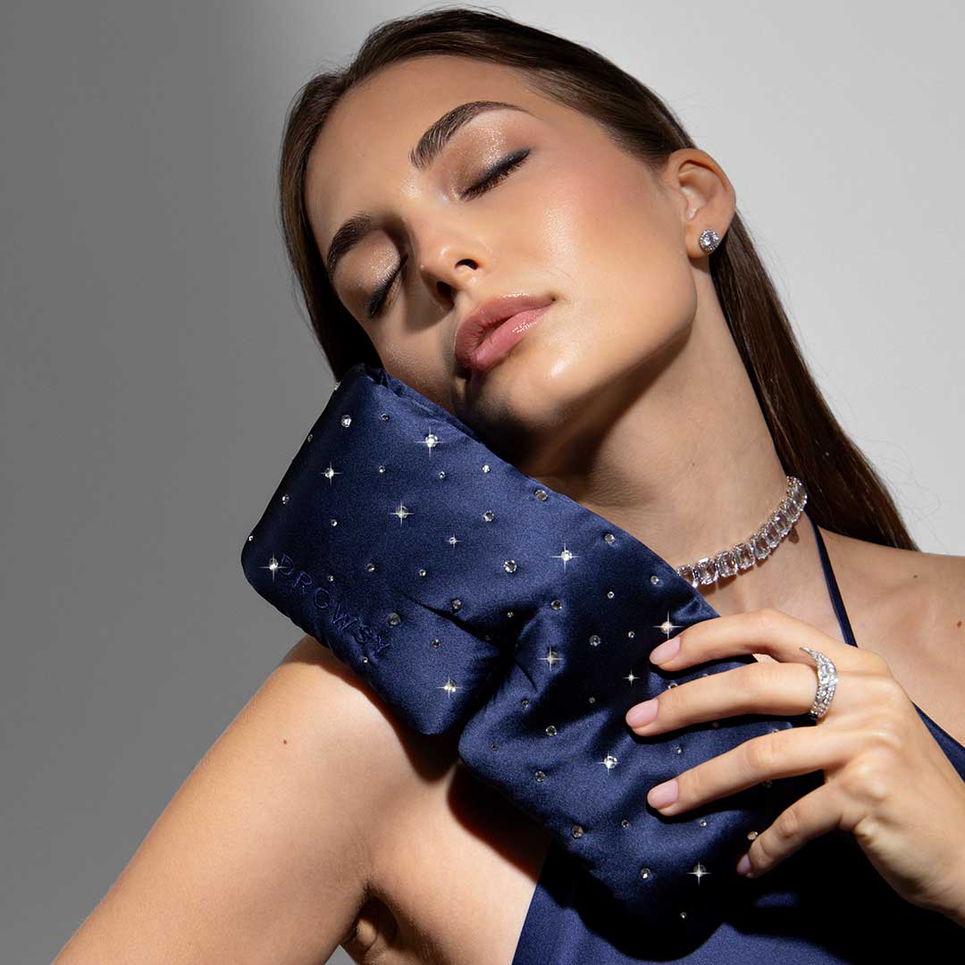 A woman with closed eyes gently holds the drowsysleepco Silk Sleep Mask Crystals by Swarovski® to her face, wearing coordinating jewelry and a dress, looking peaceful and relaxed against a neutral background.