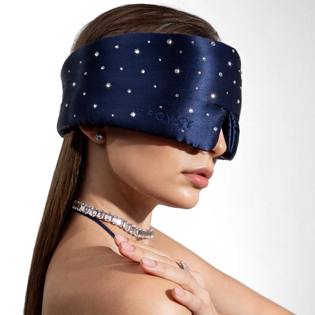 A woman models the drowsysleepco Silk Sleep Mask Crystals by Swarovski®—a navy blue blackout mask adorned with small white stars. She poses with a sparkly necklace and stud earrings against a crisp white background.