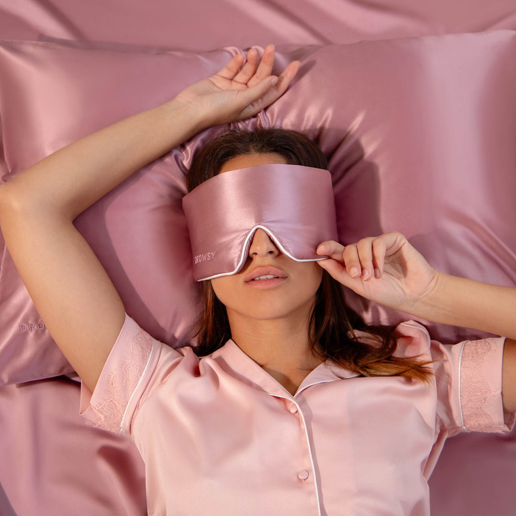 A woman relaxes on pink satin sheets, wearing drowsysleepco’s Damask Rose - Pipe Dreams Edition pajama top and adjustable sleep mask. One hand rests above her head as the other adjusts the mask over her eyes.