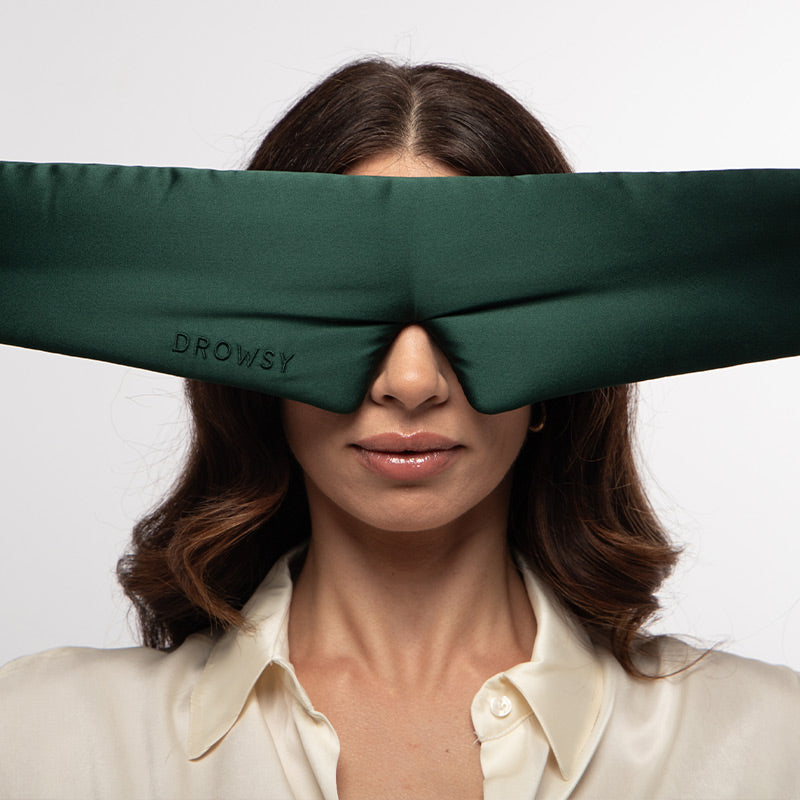 A woman wearing a cream blouse has a large, dark green padded silk sleep mask from drowsysleepco, called “Ivy Green,” covering her eyes and part of her nose against a plain white background.