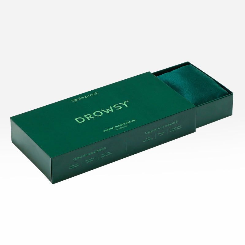 An Ivy Green sleep mask from drowsysleepco rests inside a partially open, dark green box. The minimalistic packaging features white text and product details on the lid, making it a perfect addition to your beauty sleep routine.