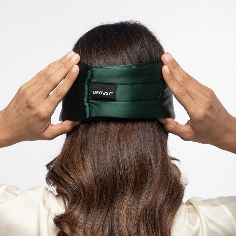 A person with long brown hair adjusts the Ivy Green padded silk sleep mask from drowsysleepco at the back of their head, shown from behind against a plain white background.