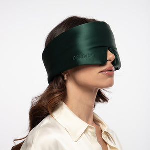 A woman with wavy brown hair is dressed in a cream-colored blouse, wearing the drowsysleepco Ivy Green padded silk sleep mask over her eyes against a plain background.