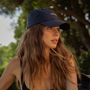 A woman with flawless hair wears the Drowsy Silk Cap in Midnight Blue by Drowsy. She is outdoors, looking to the side and enjoying the sunlight and trees—ideal for outdoor workouts.