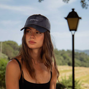 A woman with long wavy hair, in a black tank top and the Drowsy Silk Cap - Moonlight Shadow by Drowsy, sits outdoors surrounded by greenery and a vintage streetlamp—perfect for sunny days.