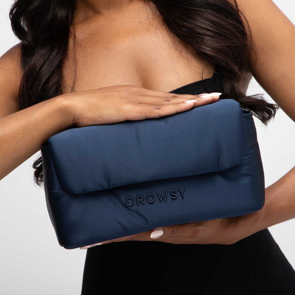 A woman in a black strapless top holds the Drowsy Sleepover bundle in Midnight Blue, featuring DROWSY embroidered on the front. Her white nails stand out, while her face remains unseen.
