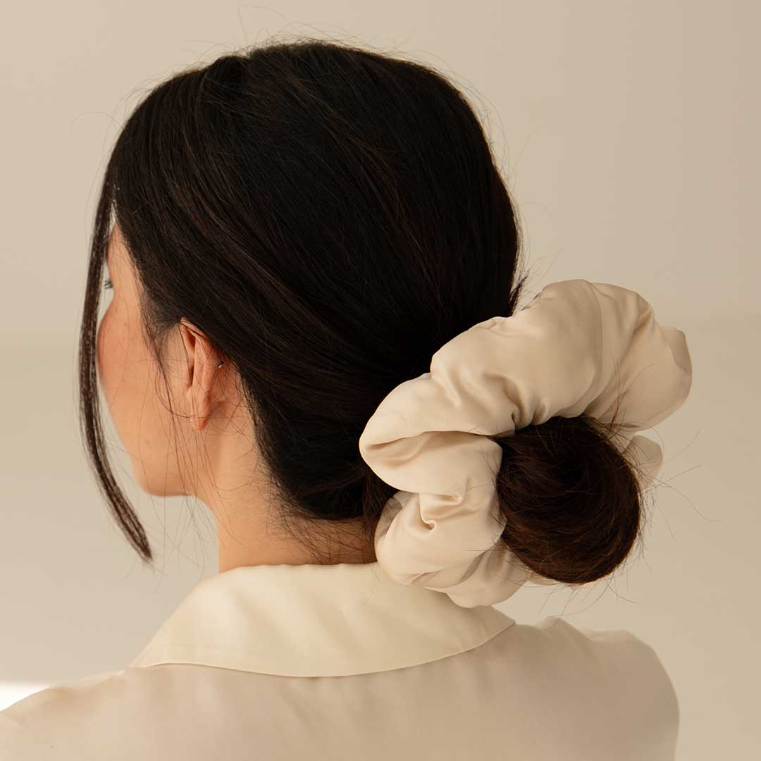 A woman with dark hair styled in a low bun, secured with the drowsysleepco Dusty Gold - Padded Scrunchie, is seen from behind wearing a cream-colored top in soft lighting.
