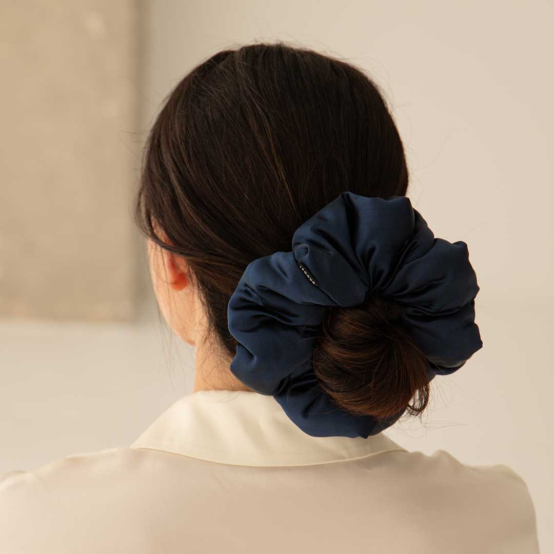 A woman with dark hair is seen from behind, wearing a cream-colored blouse. Her hair is styled in a low bun, secured with a large, dark blue satin scrunchie.