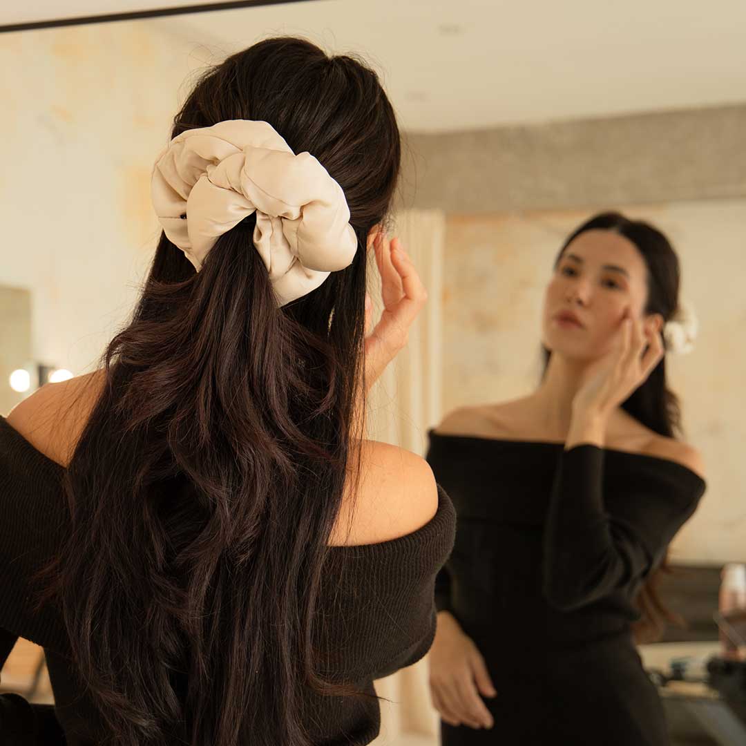 A woman with long dark hair, wearing an off-shoulder black top, looks at herself in the mirror and touches her face. Her hair is styled with the drowsysleepco Rajah Padded Scrunchie in a large cream color.