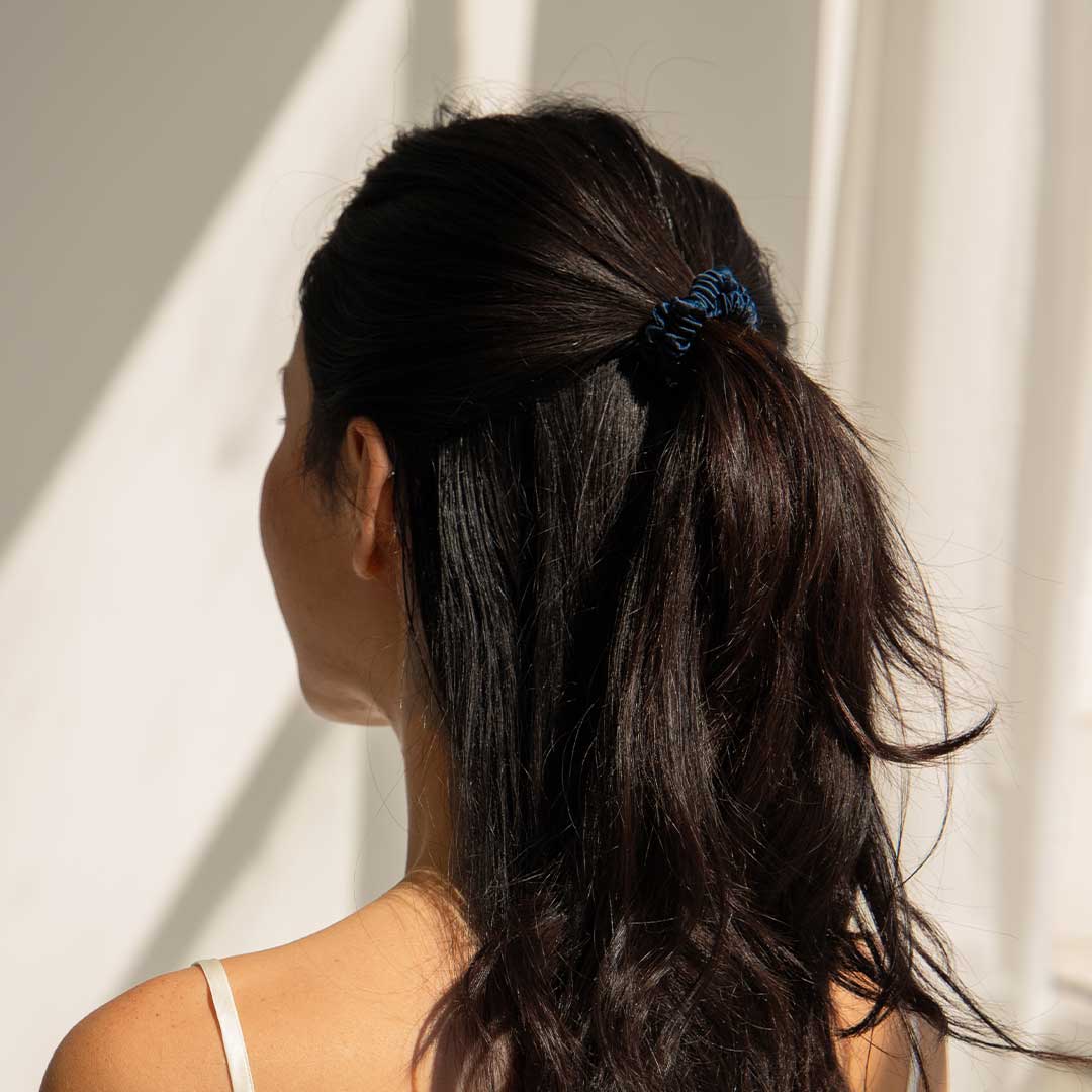 A woman with long, dark hair tied in a blue scrunchie is seen from behind, standing in front of light-colored curtains with sunlight casting soft shadows.