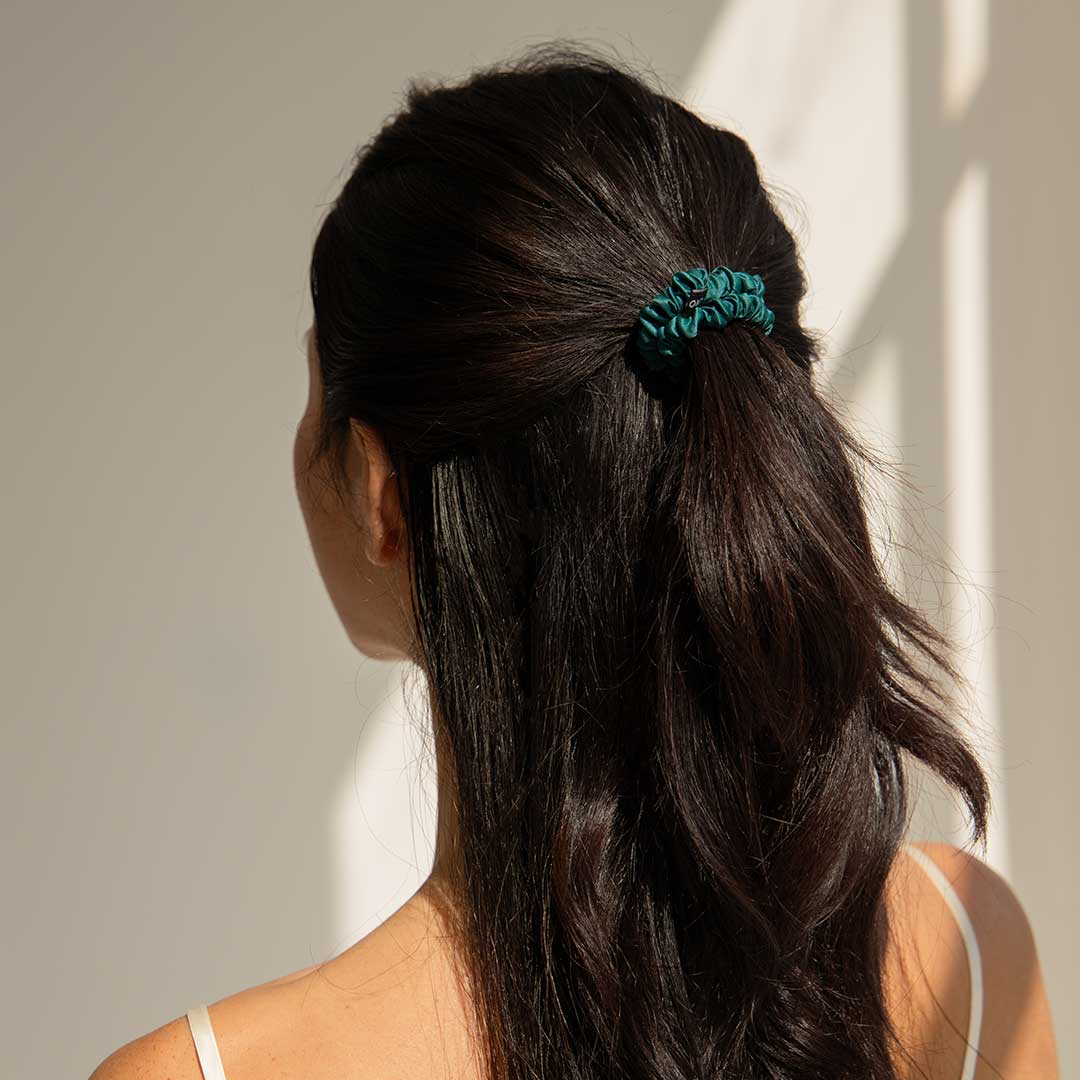 A woman with long, dark hair faces away from the camera. Her hair is partially tied back with a teal scrunchie. She is wearing a white, thin-strap top, and soft sunlight casts shadows on the wall.