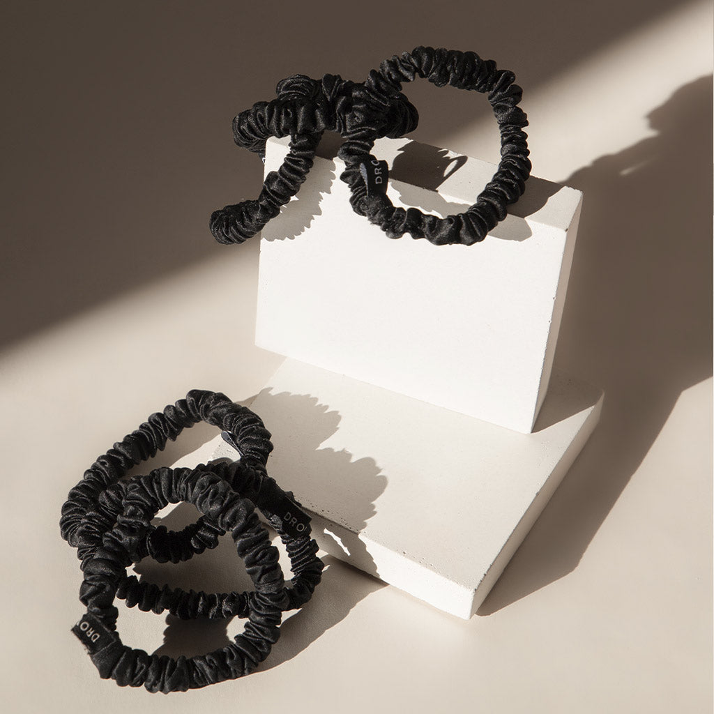Three Black Jade skinny silk scrunchies by drowsysleepco, each featuring a subtle tag, are displayed on geometric white blocks under natural light, casting soft shadows against a neutral backdrop.