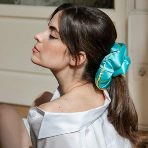 A woman with long brown hair tied back in a turquoise scrunchie relaxes indoors, eyes closed, enjoying beauty sleep on a drowsysleepco Hollywood Experience bundle - Original silk pillowcase, bathed in soft natural light.