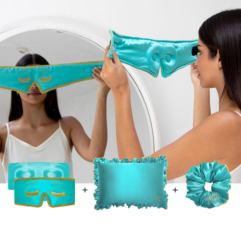 A woman in a white tank top holds drowsysleepco’s Hollywood Experience Bundle – Eyelash protecting, featuring a turquoise silk sleep mask with gold trim, matching eye mask, ruffled silk pillowcase, and coordinating scrunchie.