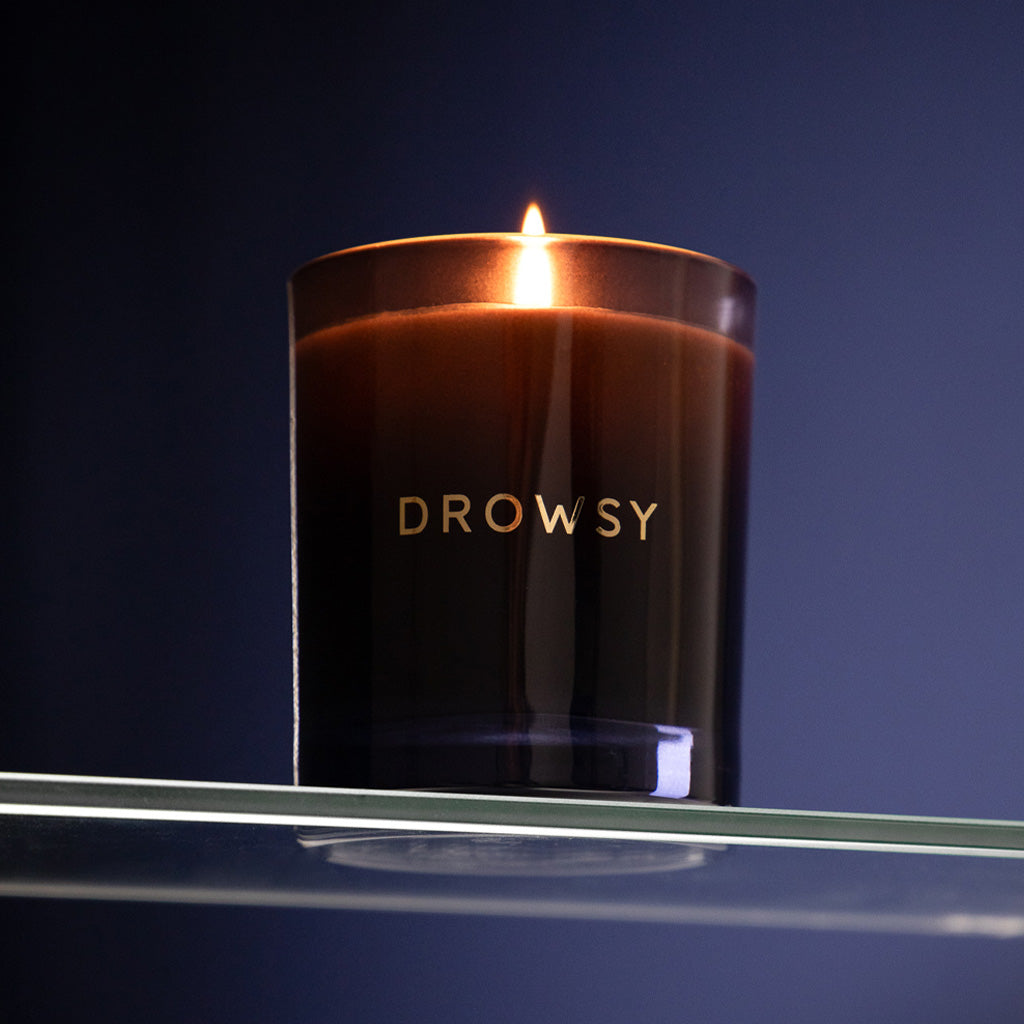 A lit black drowsysleepco Sleep Ritual Candle 220g sits on a glass shelf against a dark blue background, casting a warm glow.