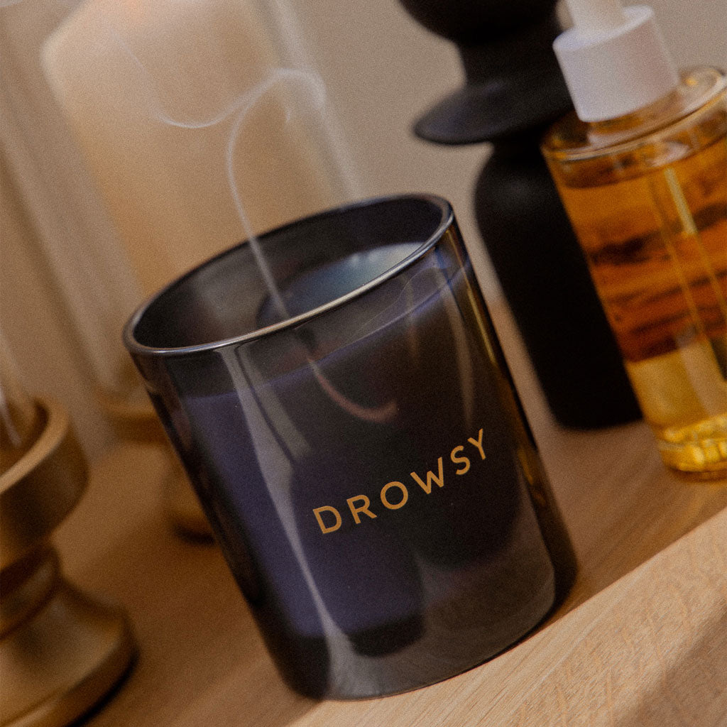 A black Sleep Ritual Candle 220g by drowsysleepco, with faint smoke rising from its wick, sits on a wooden surface surrounded by other candles and a bottle of golden natural essential oils.
