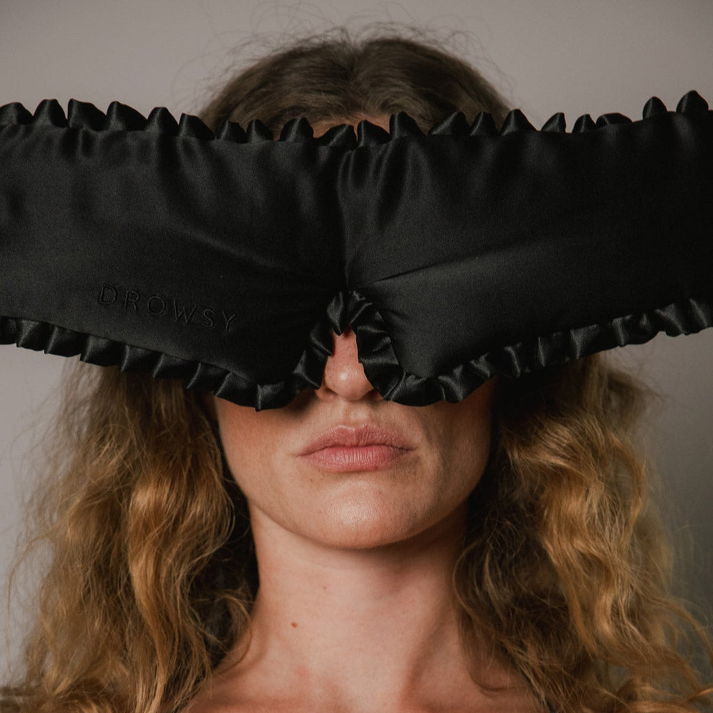 A woman with wavy blonde hair wears the drowsysleepco Manhattan Experience Bundle’s large black ruffled sleep mask, featuring faint “DROWSY” lettering, as she faces the camera against a plain background.