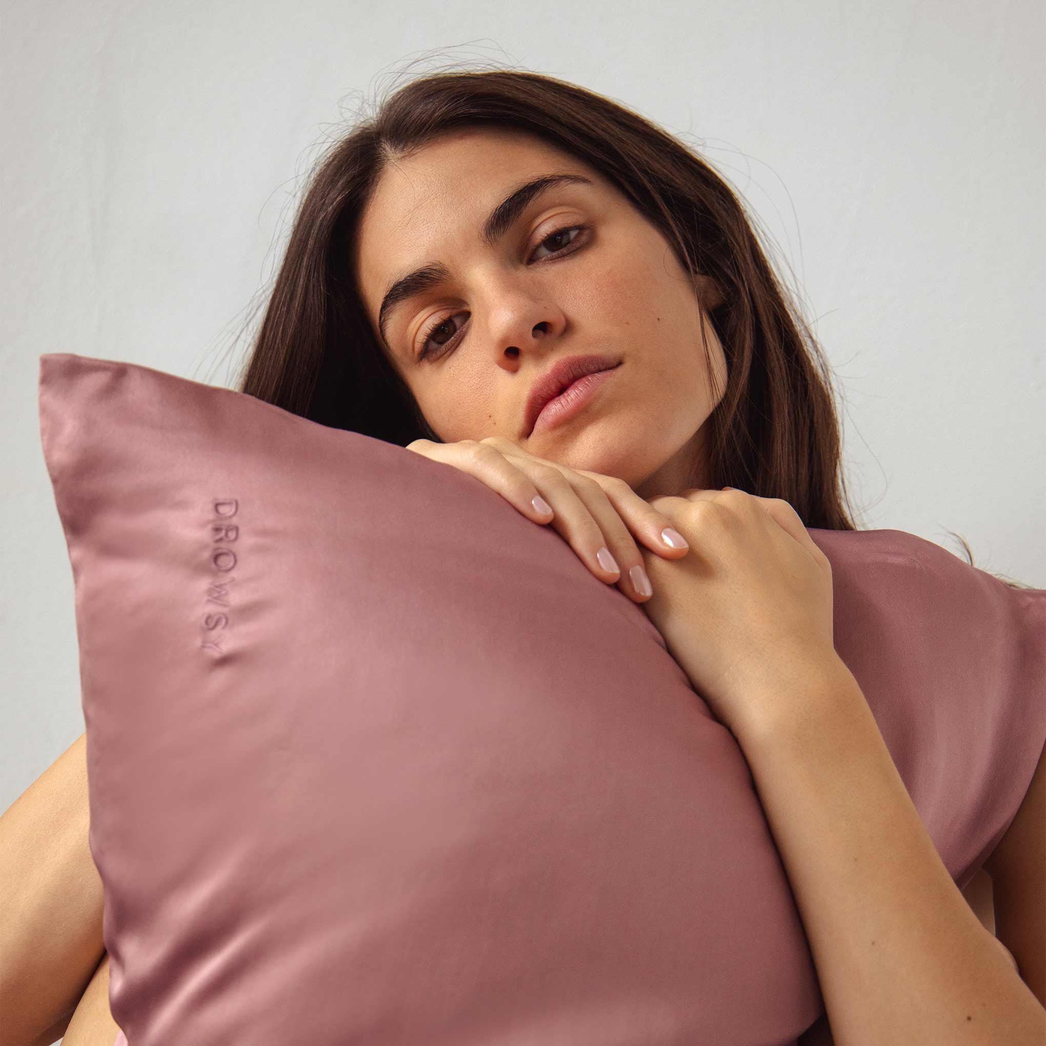 A woman with long brown hair cradles a mauve pillow embroidered with the word DROWSY, resting her chin on it and looking directly at the camera with a neutral expression.