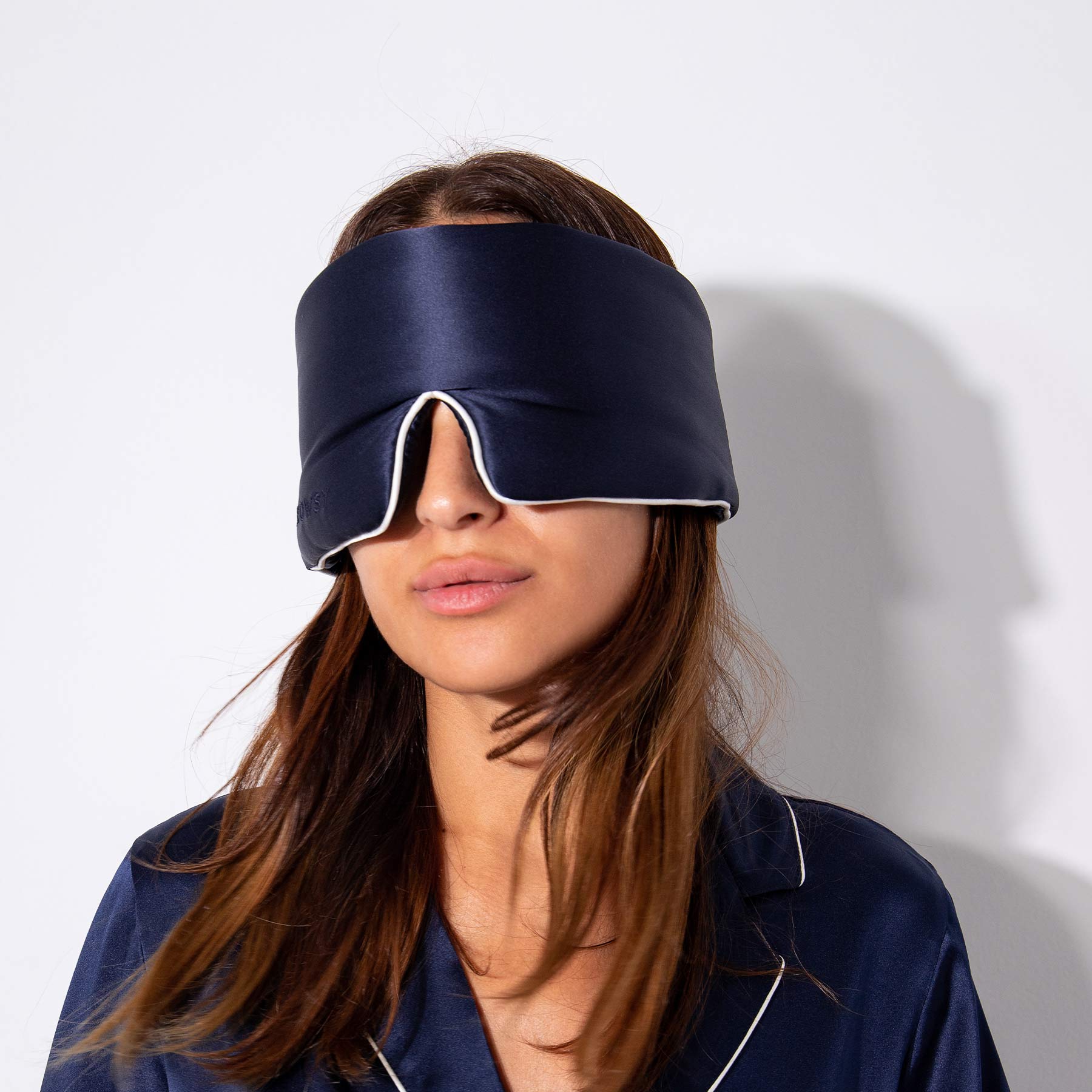 A woman wears the drowsysleepco Midnight Blue - Pipe Dreams Edition silk sleep mask and matching pajamas against a white background, her hair peeking out from beneath the adjustable mask.
