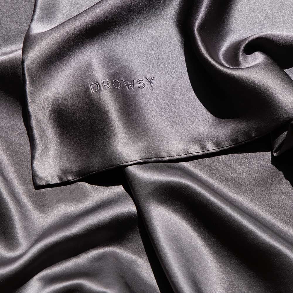 A smooth, shiny satin or silk fabric in a dark gray color, with the word DROWSY embroidered in capital letters on one corner. The material is softly draped, showcasing its glossy texture.