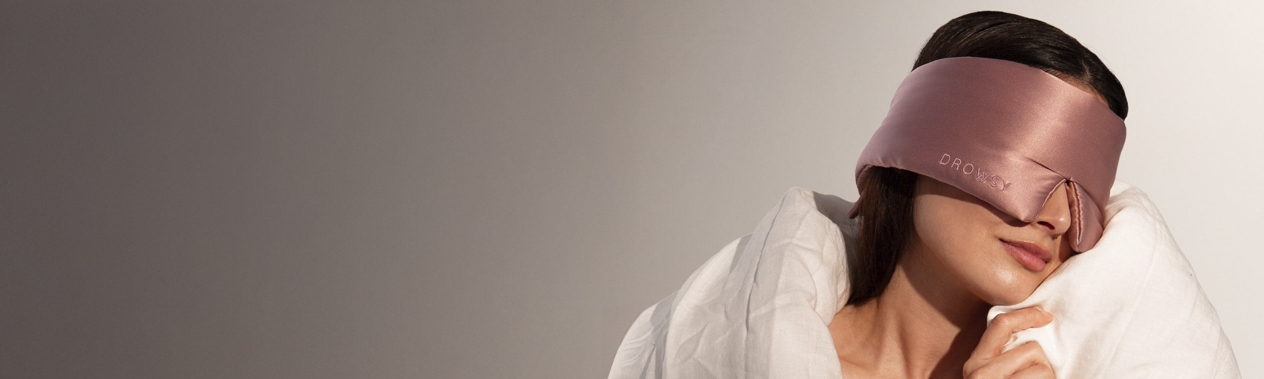 A woman wrapped in a white blanket leans her head to the side, wearing a wide, silky, rose-colored eye mask that covers her eyes and upper face. The background is a smooth, light-to-dark gradient.