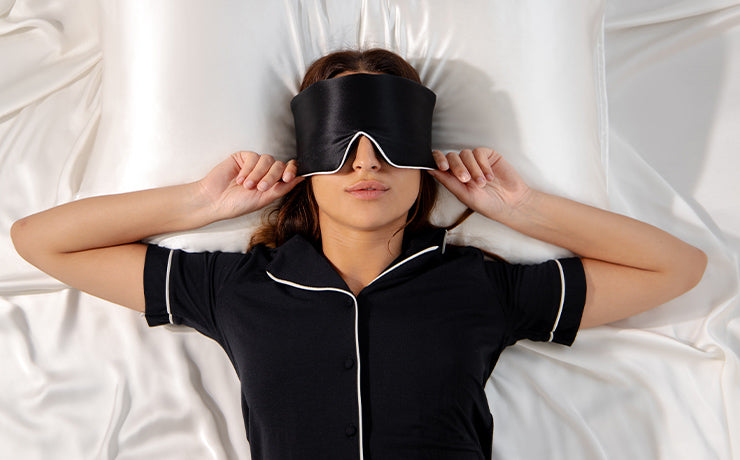 A woman lies on a white satin bed wearing black pajamas and a black sleep mask, holding the mask with both hands.
