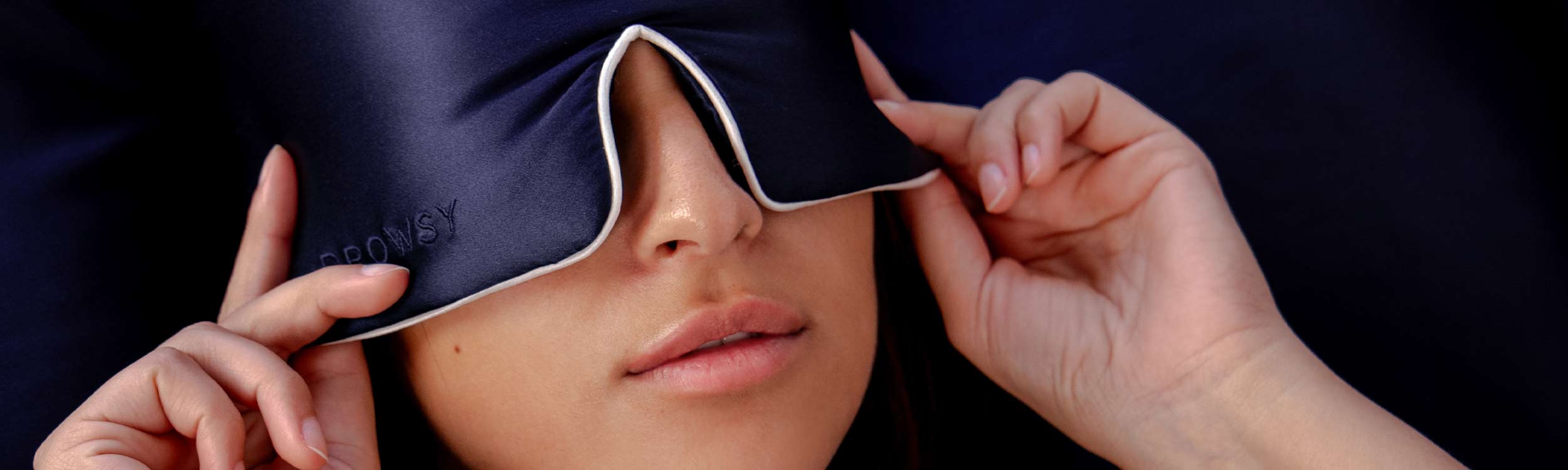 A person adjusts a dark silky sleep mask over their eyes with both hands. Only the lower half of their face is visible, showing relaxed lips and smooth skin.