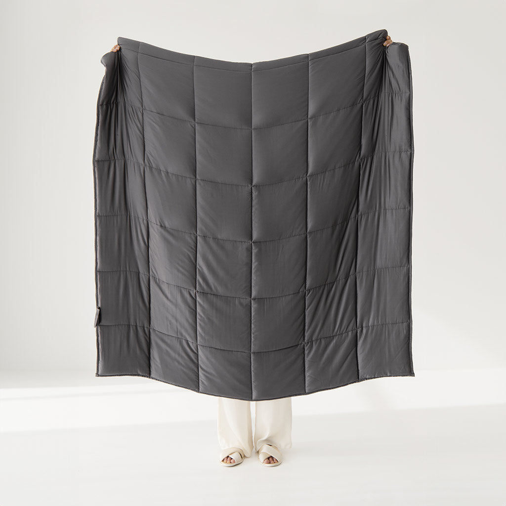 A person holds up the drowsysleepco Moonlight Shadow Silk Weighted Blanket, dark gray and large enough to cover most of their body except their feet in white pants, set against a plain light background.